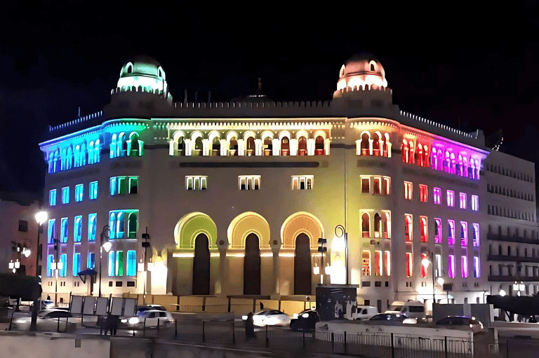 Large, light beige building at night, adorned with colorful rainbow-colored LED lights on the windows and facade, showcasing Algiers central post office