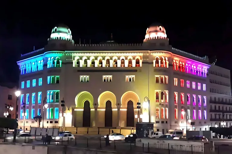 Grand bâtiment beige clair la nuit, orné de lumières LED colorées aux couleurs de l'arc-en-ciel sur les fenêtres et la façade, mettant en valeur la poste centrale d'Alger