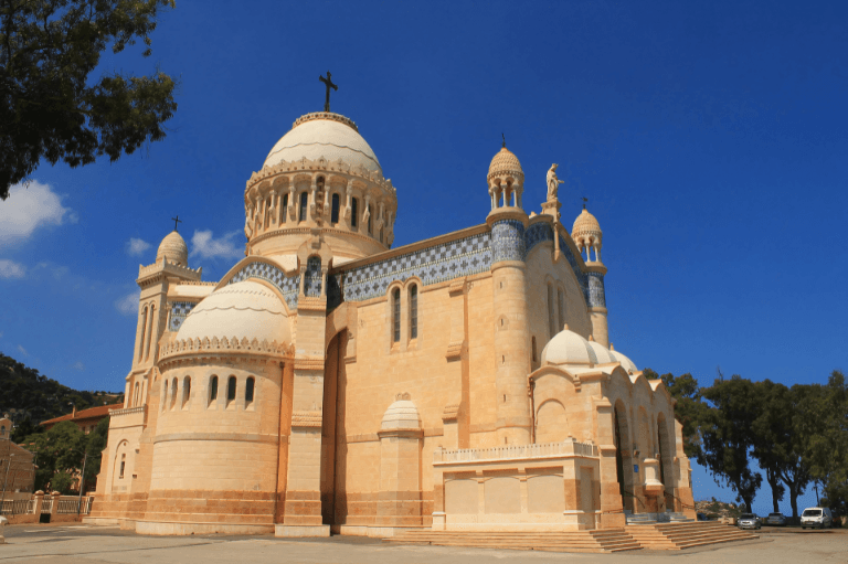 Exterior view of Notre-Dame d'Afrique Basilica in Algiers, Algeria, showcasing its neo-Byzantine architecture with large domes and intricate details against a clear blue sky.
