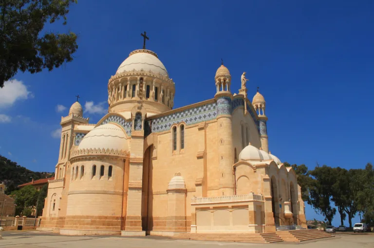 Vue extérieure de la basilique Notre-Dame d'Afrique à Alger, en Algérie, présentant son architecture néo-byzantine avec de grands dômes et des détails complexes sur un ciel bleu clair.