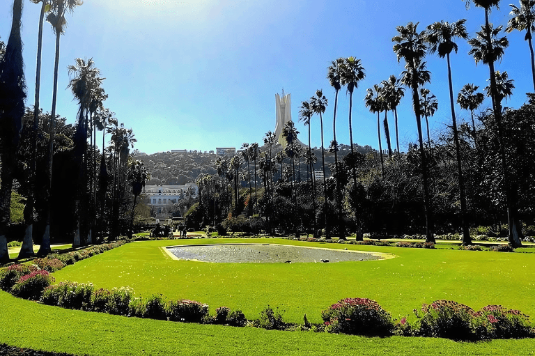 A Botanical Garden Hamma algiers park with manicured lawns, flowerbeds, palm trees, and a reflecting pool; a martyrs memorial monument is visible in the background.