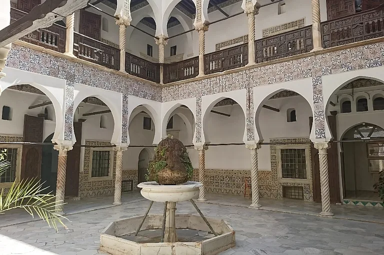 Cour historique de style ottoman avec des arches ornées, des carreaux complexes et une fontaine centrale en pierre, mettant en valeur l'architecture traditionnelle et les éléments de design de la casbah d'Alger