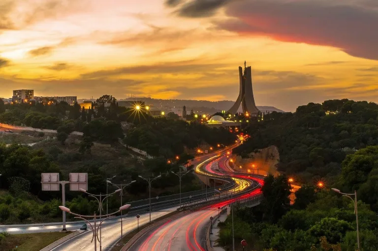 Autoroute serpentant à travers un paysage avec un important mémorial des martyrs d'Alger au coucher du soleil, illuminé par les lumières de la ville.