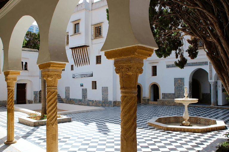 A courtyard with black and white checkered tiles is surrounded by white buildings featuring arched columns with intricate carvings. A small fountain graces the center, while lush trees provide shade, complementing the serene architectural setting of the bardo national meseum of algiers