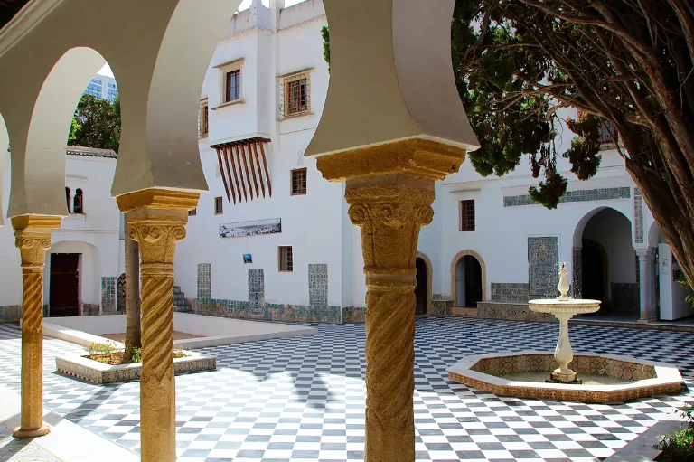 Une cour aux carreaux à damiers noirs et blancs est entourée de bâtiments blancs aux colonnes arquées aux sculptures complexes. Une petite fontaine orne le centre, tandis que des arbres luxuriants fournissent de l'ombre, complétant le cadre architectural serein du musée national du Bardo d'Alger