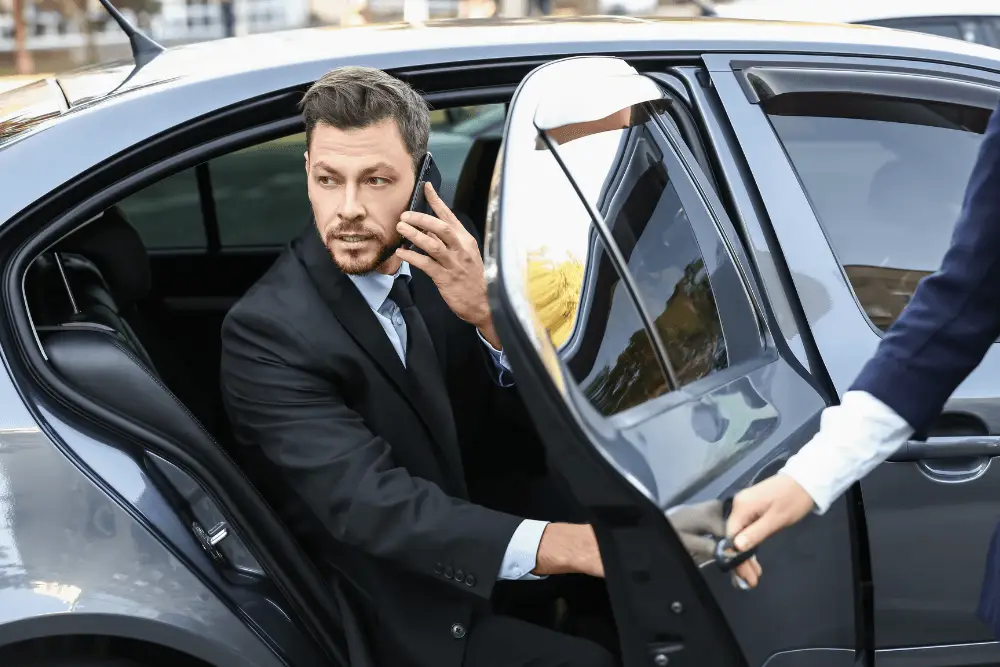 Businessman in a suit talking on a cell phone while stepping out of car with assistance from a private driver.