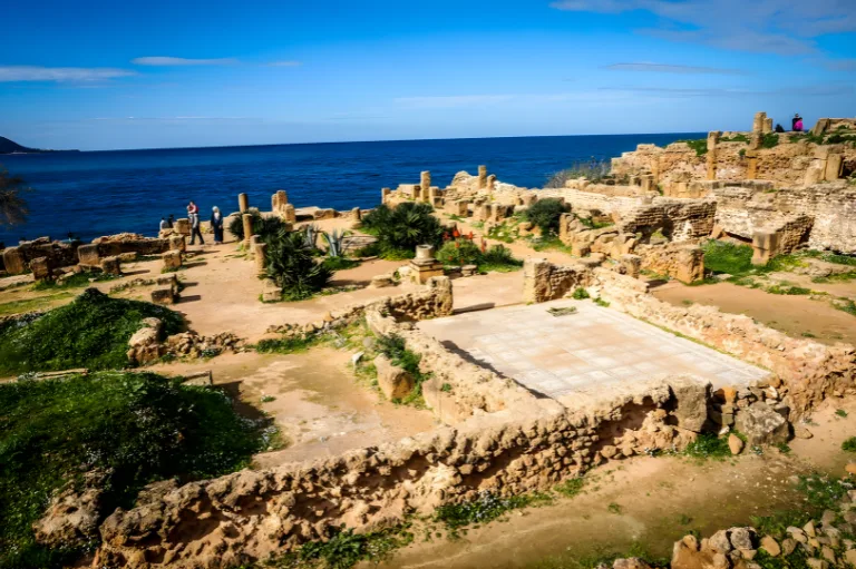 Les ruines romaines antiques de Tipaza surplombent la mer Méditerranée d'un bleu profond sous un ciel d'un bleu éclatant, tandis que des touristes explorent le site.