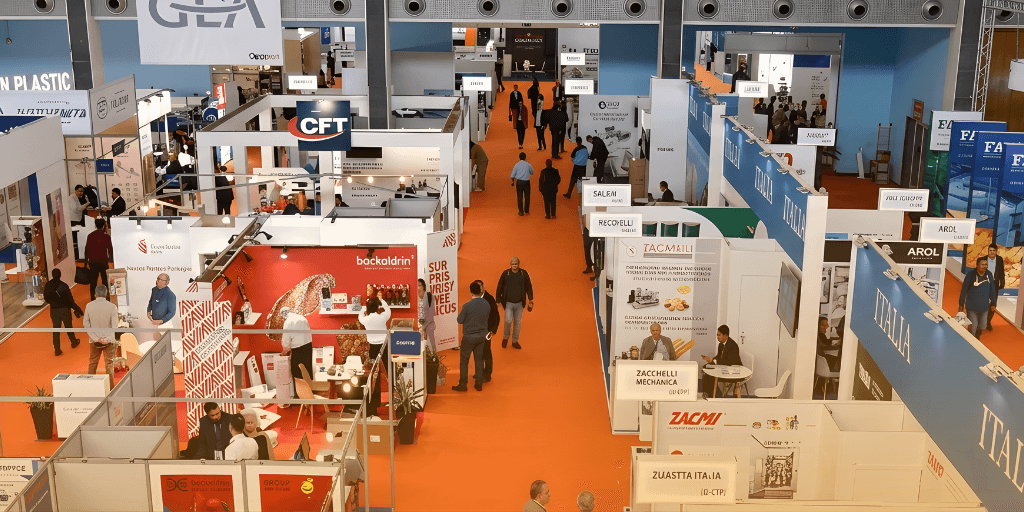Busy DJAZAGRO Trade Fair floor with attendees walking along an orange carpet between numerous white and branded exhibition booths.