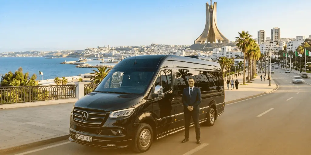 Guía turística de Argelia: Un chófer se encuentra junto a una furgoneta Mercedes-Benz Sprinter negra en una carretera costera de Argel, con el monumento Maqam Echahid visible al fondo.